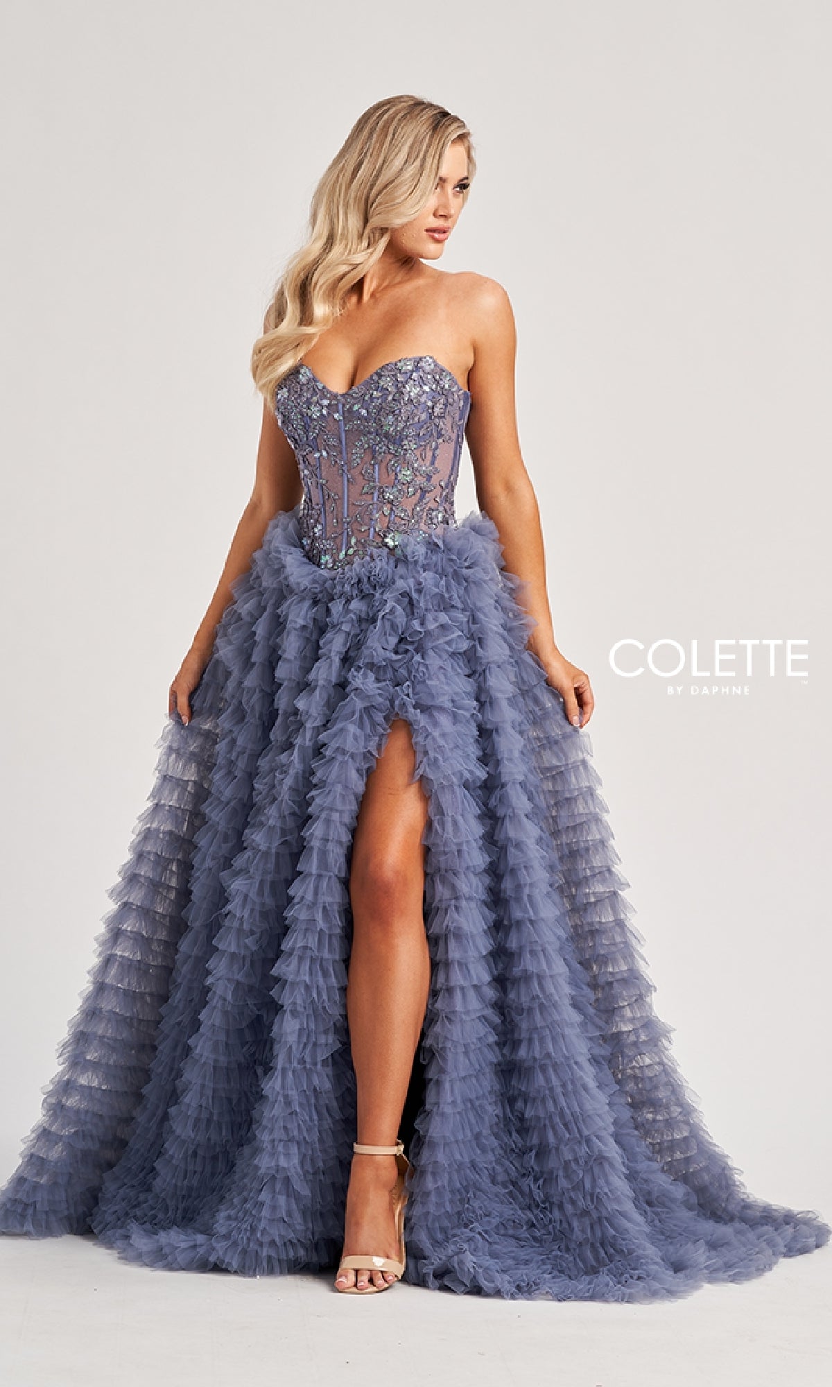 A woman poses against a plain background wearing the Colette CL8170 long prom dress—a strapless, beaded lace corset ballgown with a layered tulle skirt and high slit.